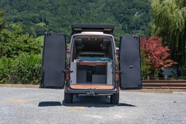Ein Blick in den VW Crafter 4x4 mit geöffneten Hecktüren. Der Camper Van von Cocoon Vans verfügt über ein Hubbett, welches auf dem Bild heruntergelassen ist.