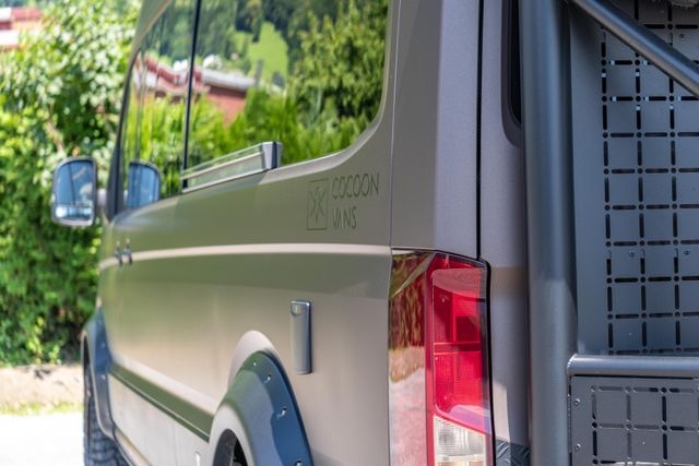 Nahaufnahme der Seite eines VW Crafters 4x4 mit dem Cocoon Vans Logo. Grünes Laub und bunte Blumen spiegeln sich im Fenster.