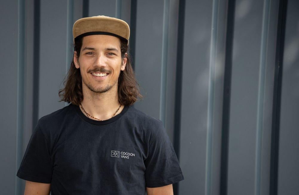 Ein Mann steht vor einer grauen Wand. Auf dem Kopf ein baiges Camp, mitellange Haare und einen Schnurrbart. Der Mann trägt ein Cocoon Vans T-Shirt. Es ist Sommer