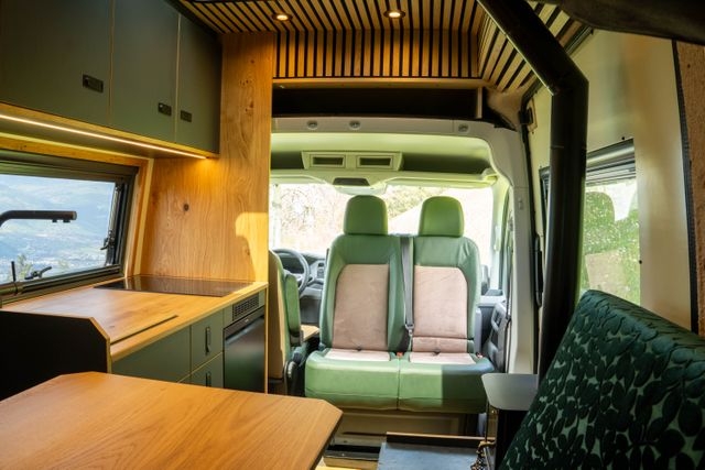 Grossansicht des Innenbereichs vom MAN Camper Vans. Die Doppeldresitzkonsole ist gedreht. Das Spühlbecken ist abgedeckt. Zu sehen ist auch der Tiny Stove und die Lehne der Sitzecke aus einem samtgrünen Stoff sowie einen Teil des Hubtischs.