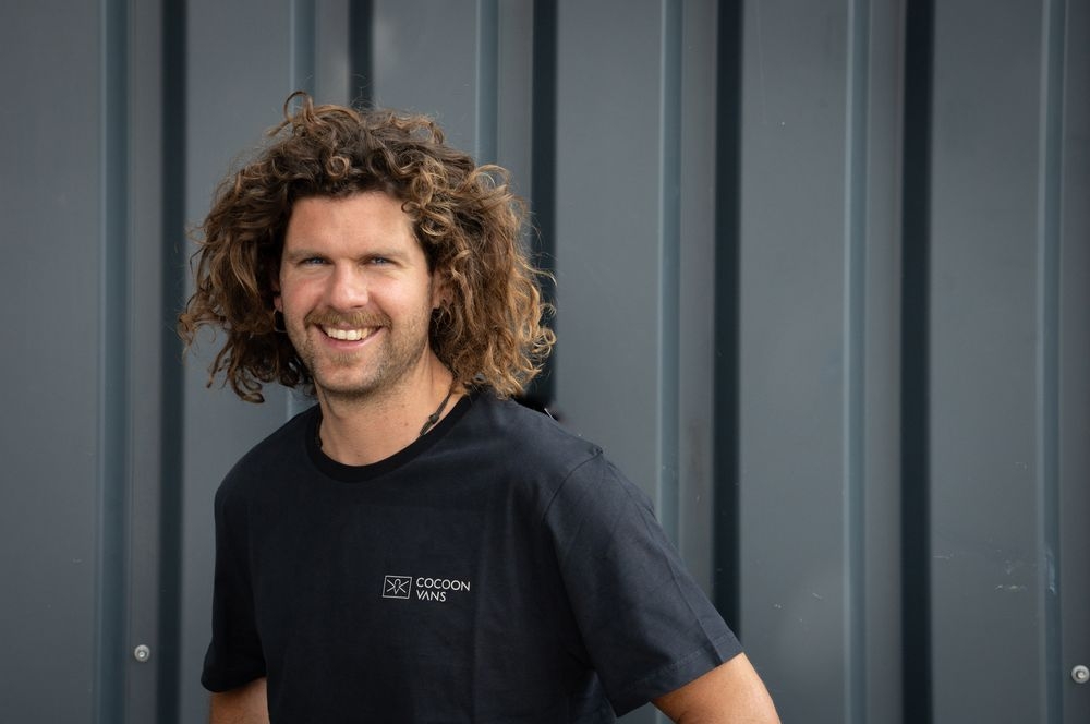 Adi steht im freien und hat im rechten Ohr ein grösseres Ohring. Der Hintergrund ist aus dunklem Wellblech